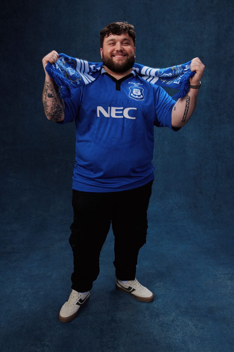 Smiling bearded man wearing a vintage Everton FC blue NEC-sponsored football shirt, holding a blue and white Everton scarf above his head, with tattoos visible on